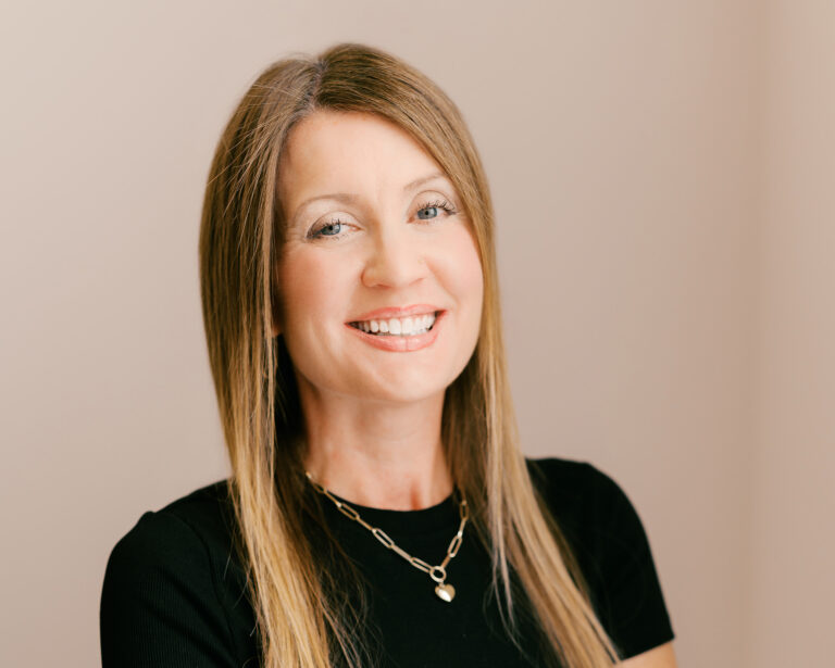 Brooke, a white female with long dirty blonde hair, poses and smiles with a black tee and gold necklase