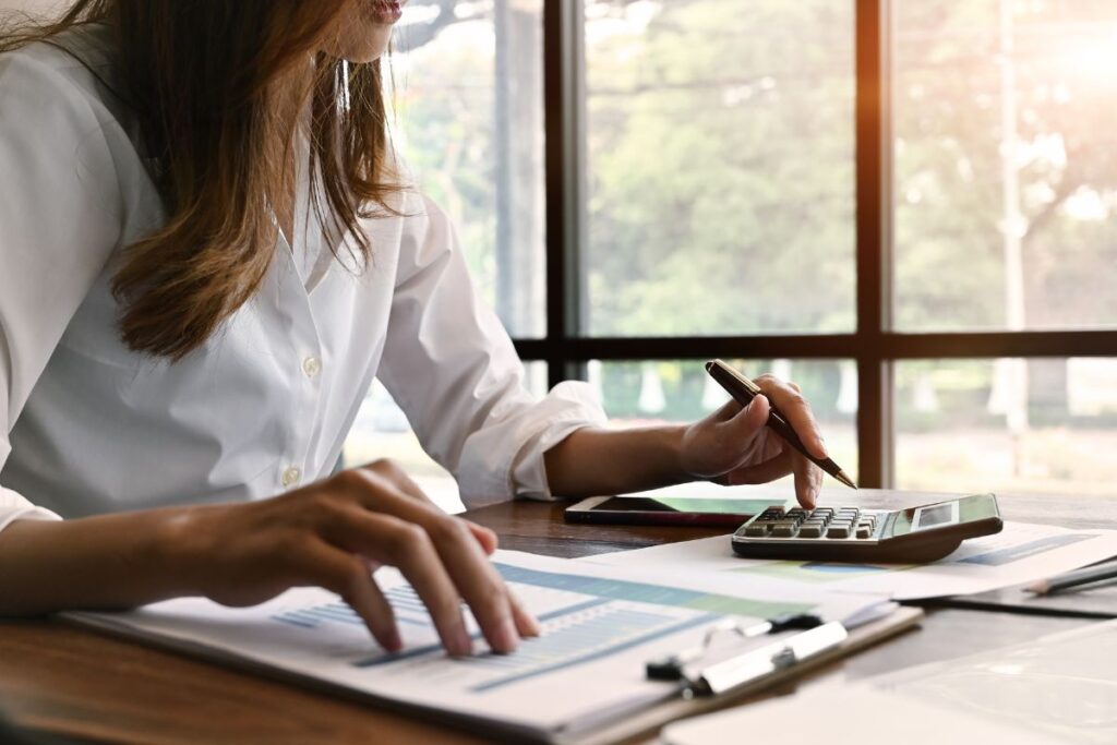 Woman with brown hair and a white button up sitting at a desk calculating finances.